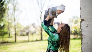 Mother joyfully lifts her baby outdoors in a bright, sunny setting.